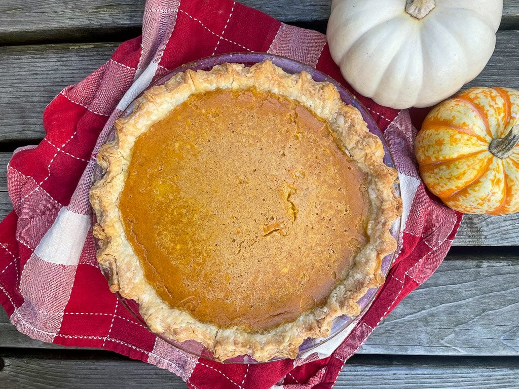 A pumpkin pie on a checkered tea towel surrounded by small pumpkins.